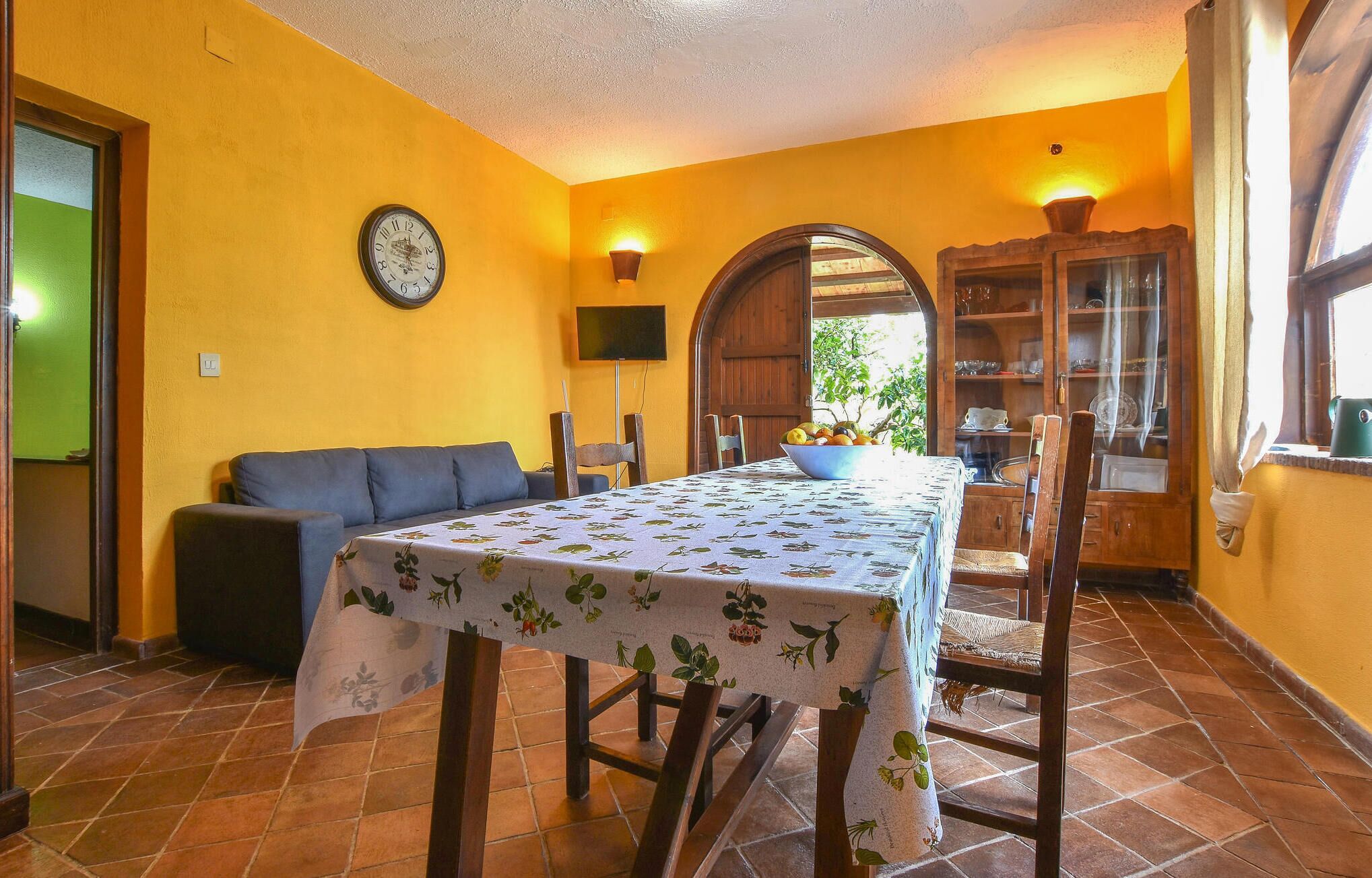 A dining area with a table set for two, a sofa, a clock, and a wooden cabinet with glass doors.