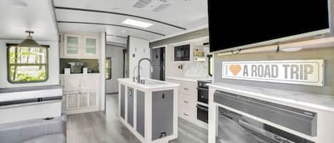 A modern RV interior with a kitchenette, a dining area, and a TV mounted on the wall.