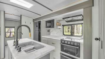 A compact RV kitchen with a sink, stove, microwave, and a window with a view of greenery.