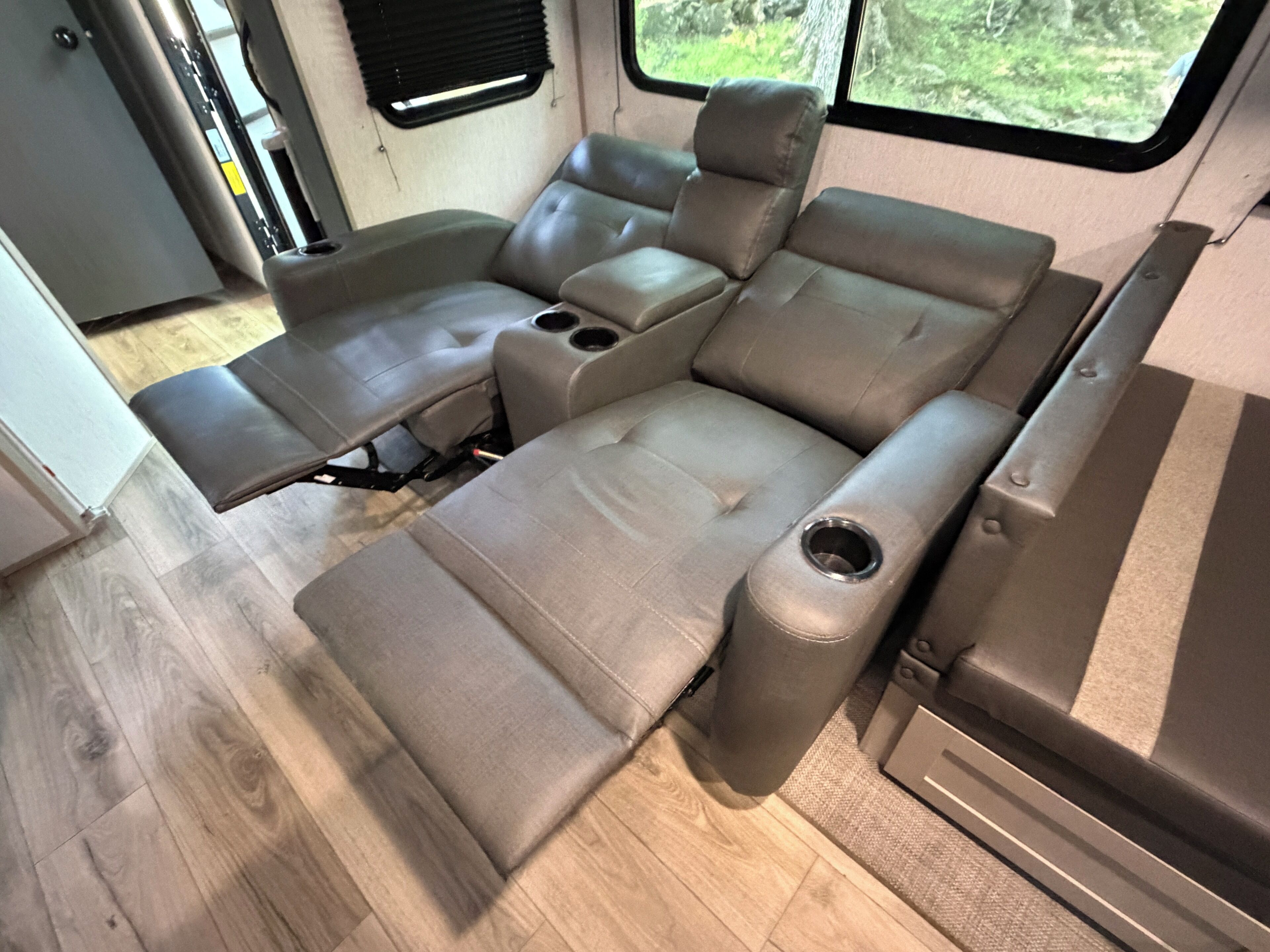 A compact RV interior with a sectional sofa, a small table, and a window providing a view of greenery.