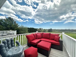 A covered outdoor deck with a sectional sofa and ottomans, a stone sculpture, and a view of a green landscape. - Tradewind Haven II - 5Bed Townhouse Near Newport! (Middletown)