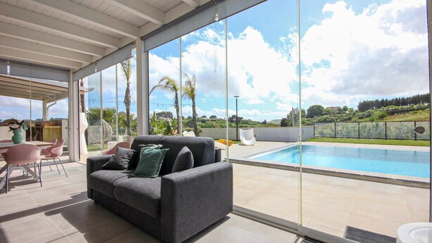 A modern living room with a grey sofa, a dining table with chairs, and a large window overlooking a swimming pool. - Amazing home in Salemi (Salemi)
