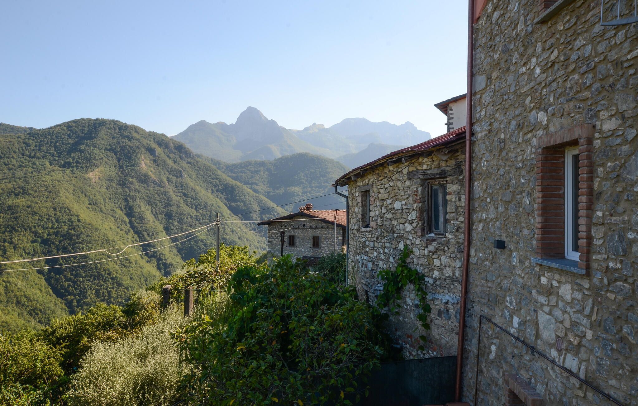 Un edificio in pietra con tetto in tegole rosse, immerso nel verde e con montagne sullo sfondo.