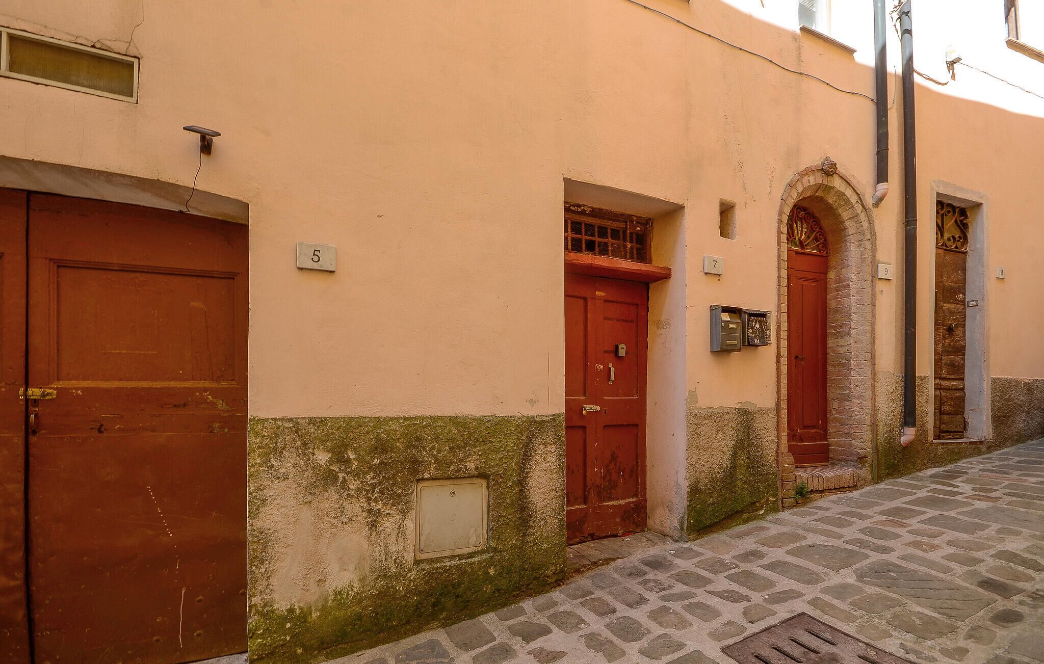 Una estrecha calle adoquinada con un edificio que tiene dos puertas rojas, un ventanal pequeño y un buzón.
