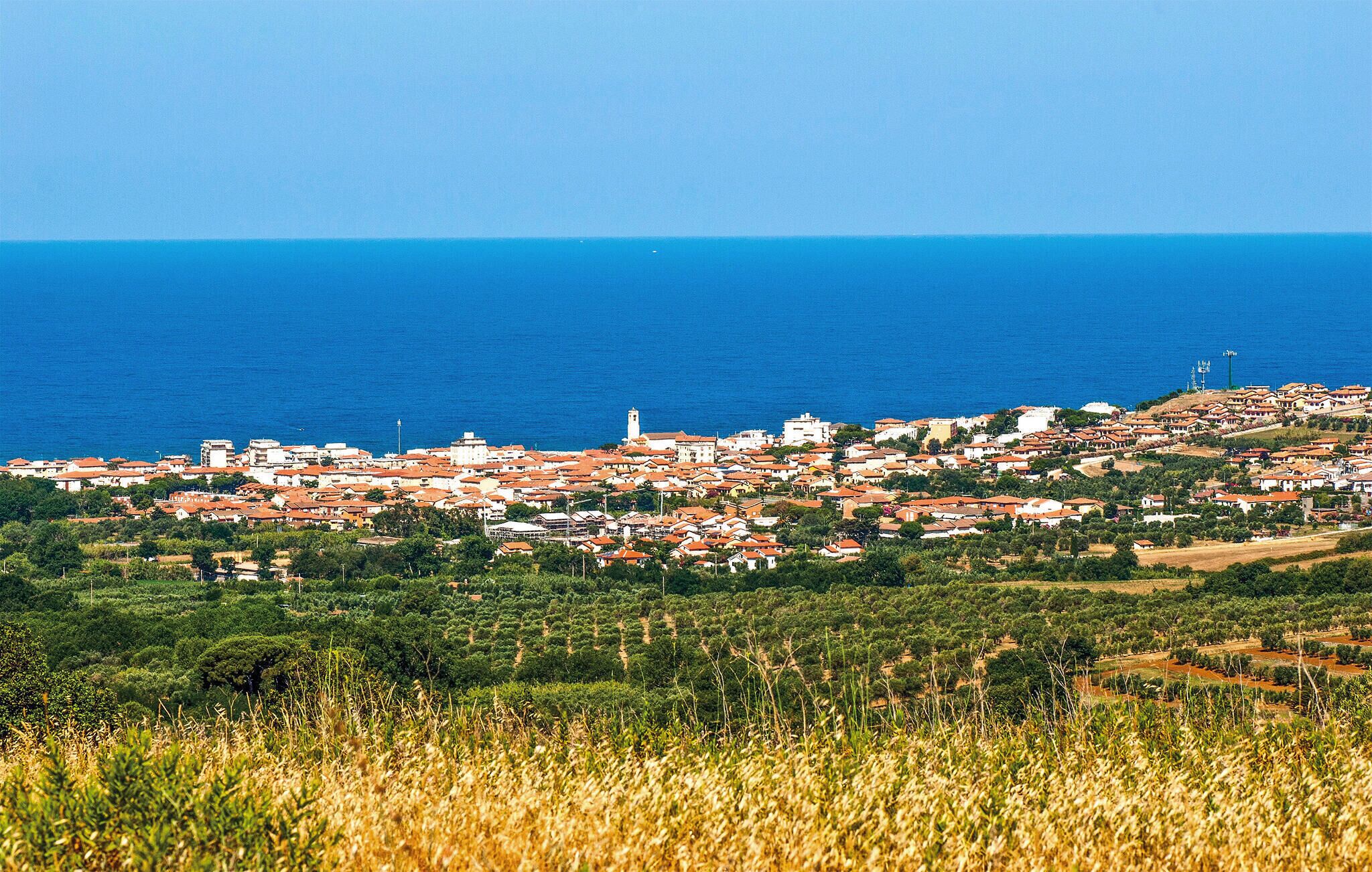 A coastal town with a mix of residential buildings and greenery, a prominent white tower, and a clear view of the sea.