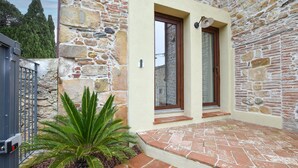 Un edificio de piedra con una puerta y una ventana, un pequeño jardín con una planta de palmera y un acceso pavimentado.