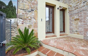 Ein Steinhaus mit einer Tür und einem Fenster, ein kleiner Garten mit einer Palme und einem gepflasterten Eingang.