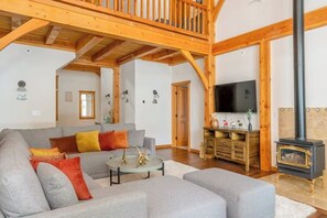 Ein geräumiges Wohnzimmer mit hoher Holzbalkendecke, einem grauen Sofa, einem Couchtisch, einem Fernseher und einem Kaminofen.