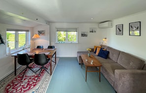 Ein Wohnzimmer mit Essbereich, einer Couch, einem Couchtisch, einem Teppich und einem Fenster mit Blick ins Grüne.