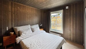 Una camera da letto con soffitto in legno, una grande finestra, un letto con lenzuola a righe, una lampada da comodino e un tavolino di servizio.