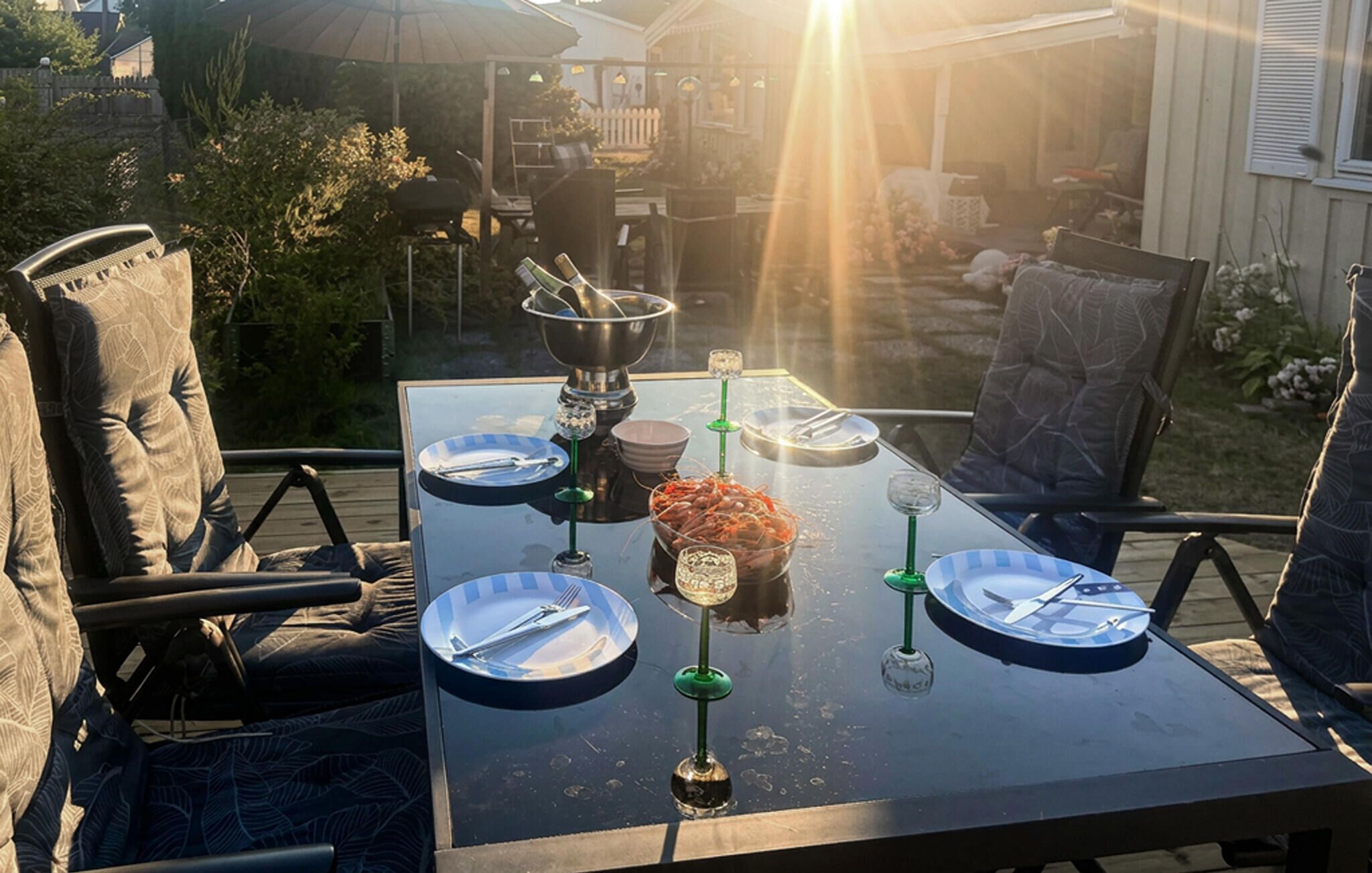 An outdoor dining table set with plates, cutlery, and a bowl of food, surrounded by chairs and a grill.