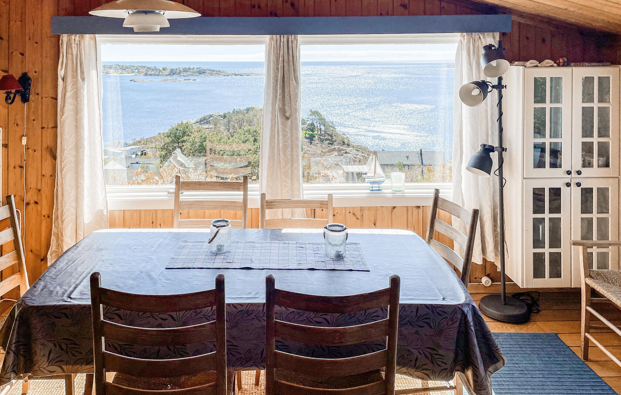 A wooden interior with a table and chairs, a view of the sea, and a cabinet with glass doors.