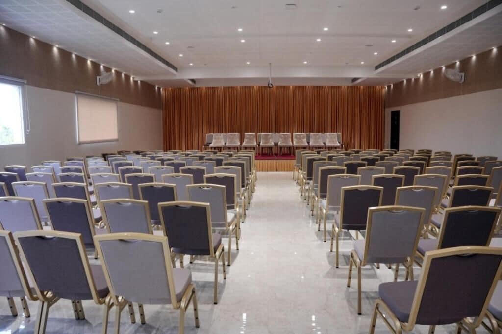 Un salón de conferencias con filas de sillas, un escenario y una gran pantalla.