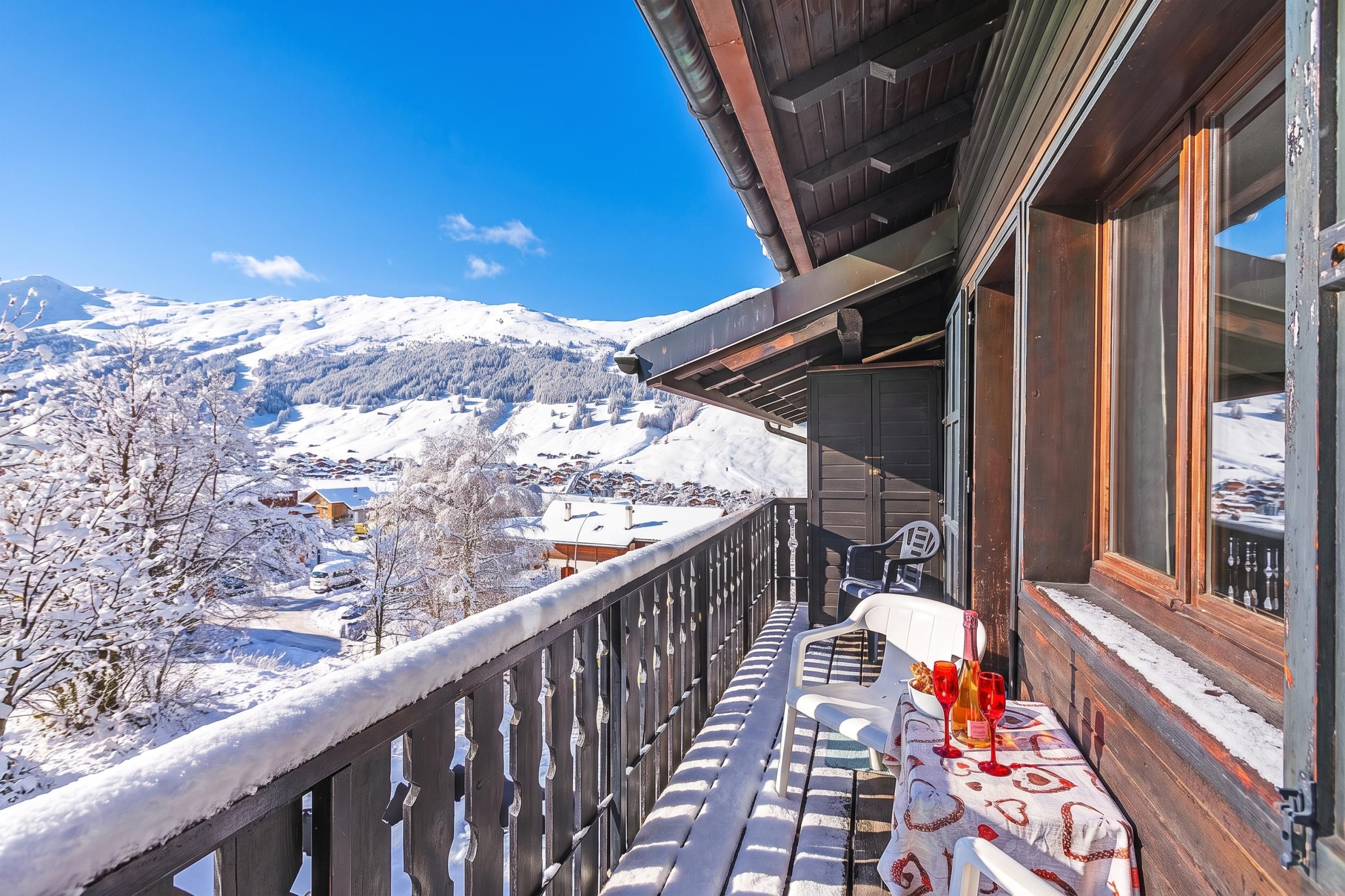 Ein Balkon mit Blick auf schneebedeckte Berge, einem Holgitter und einem Tisch mit Gläsern und einer Flasche.