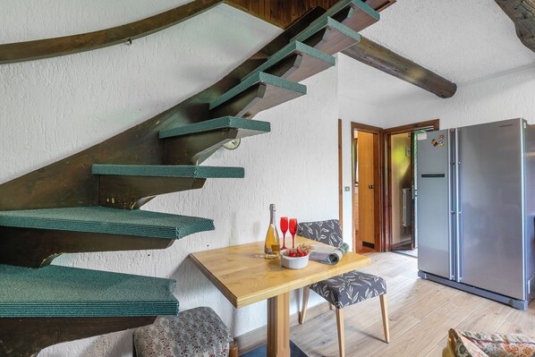 Un escalier aux marches vertes, une table en bois sur laquelle se trouve un saladier rempli de fruits et deux verres rouges, une chaise à motifs et un réfrigérateur, le tout dans une pièce aux poutres apparentes et aux murs blancs.
