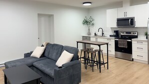Moderni keittiö, jossa on valkoiset kaapit, mikroaaltouuni, uuni ja kahvinkeitin. Olohuoneessa on harmaa sohva, jossa on koristetyynyjä, sekä pieni pöytä ja jakkarat.