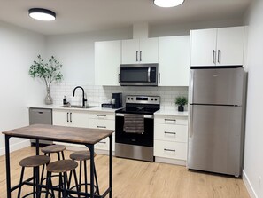 A modern kitchen with white cabinets, a stainless steel refrigerator, a microwave, an oven, and a coffee maker. There is a wooden table with stools and a potted plant on the counter. - Upscale 1BR Suite in New Build | Nature Escape (Kelowna)