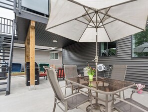 An outdoor patio with a glass table and chairs, a large umbrella, and a grill. - Modern 5BR Home Near Lions Gate Glass Deck BBQ (West Vancouver)