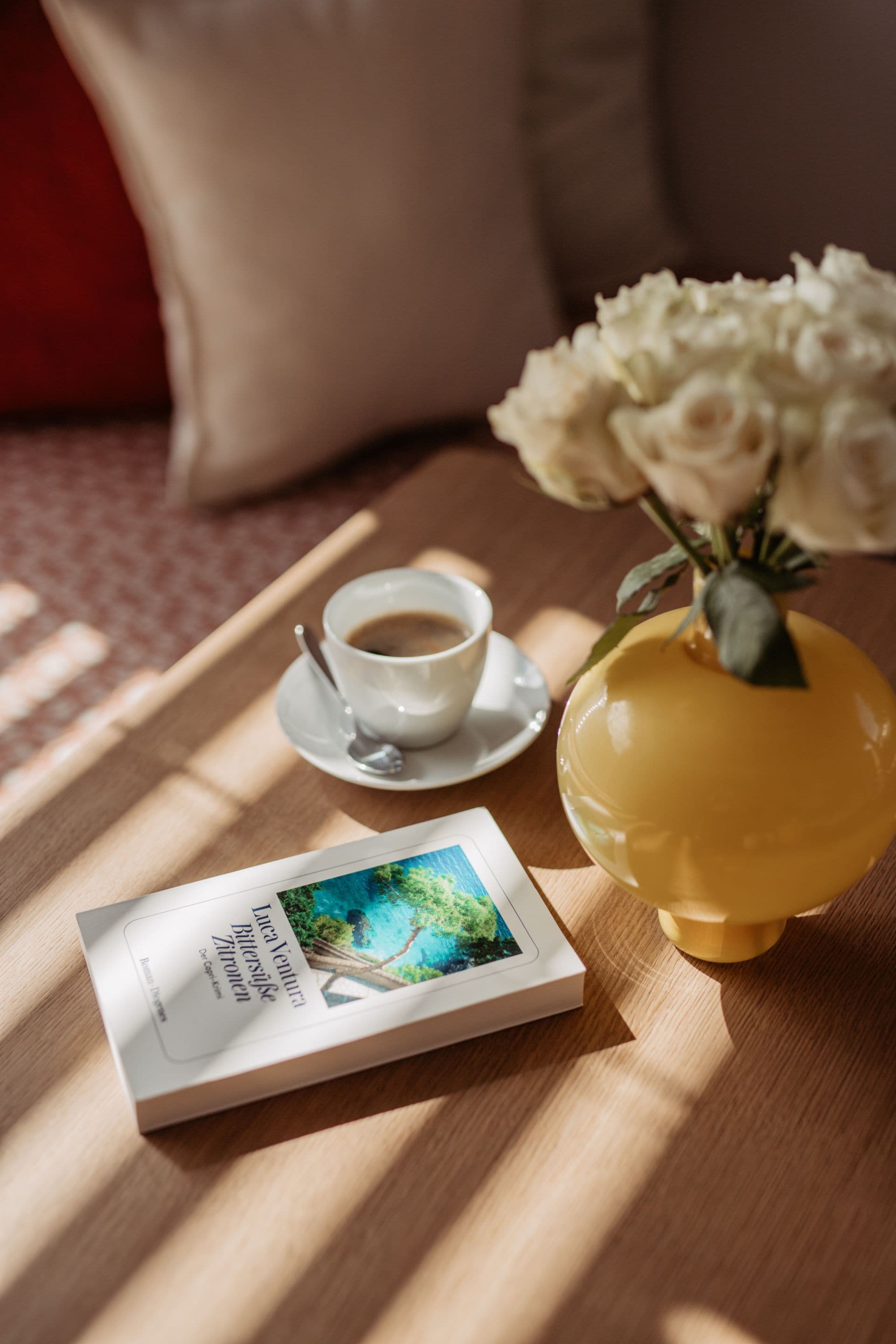 A book, a cup of coffee, and a vase with flowers on a wooden table.