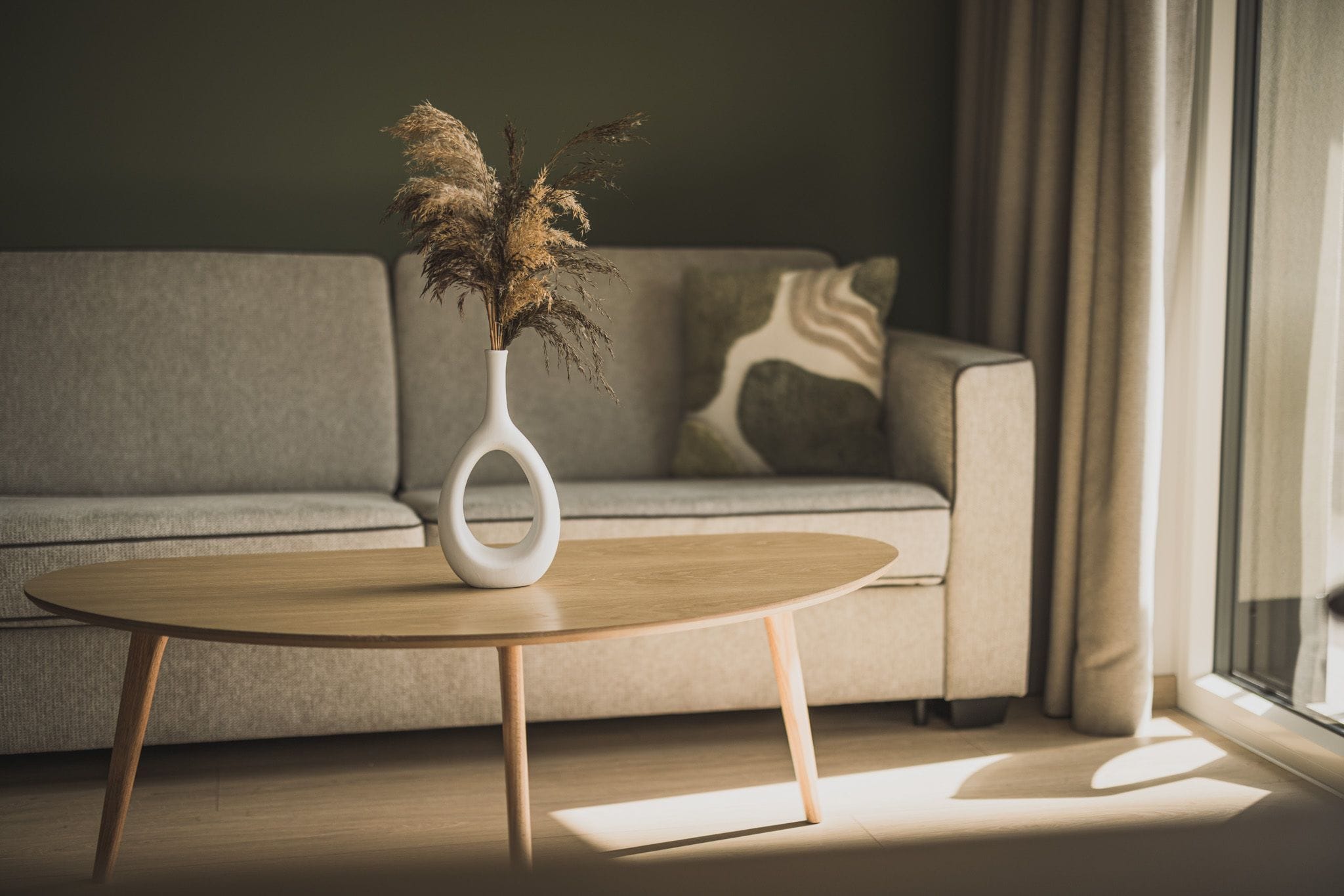 A modern living room with a round wooden coffee table, a white vase with dried plants, a grey sofa, and a patterned throw pillow.