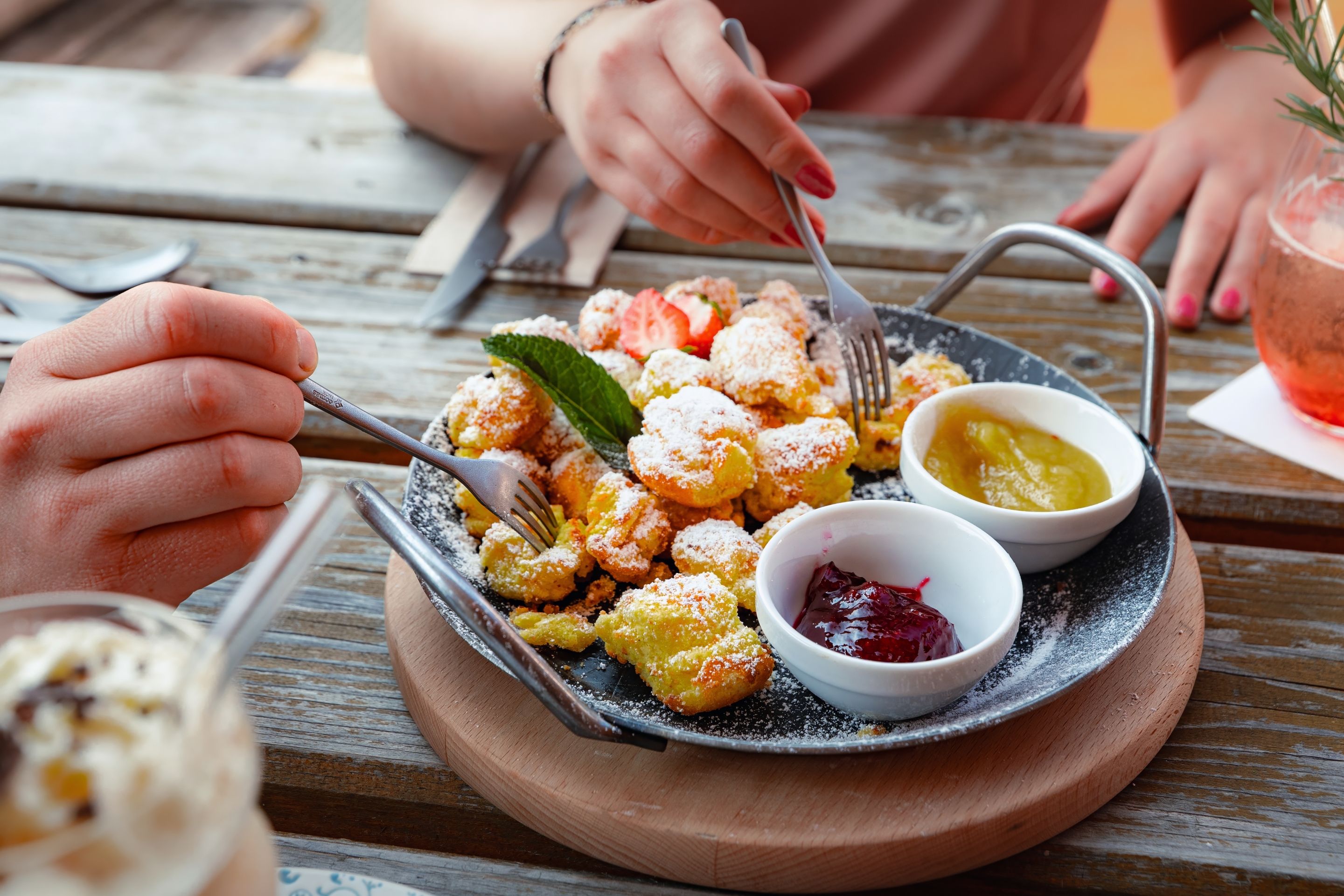 A wooden table with a plate of fritters, two small bowls of jam, and a glass of iced drink.