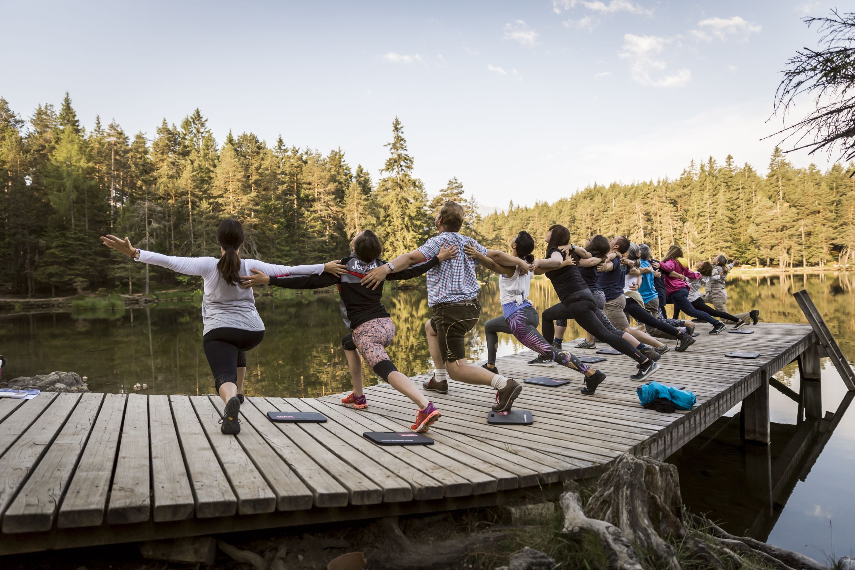 Un grupo de personas haciendo ejercicio en un muelle de madera junto a un lago, con un bosque frondoso al fondo.