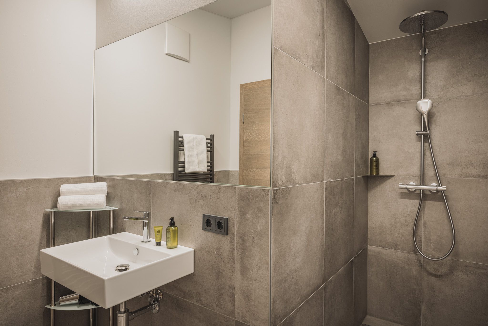 Ein modernes Badezimmer mit einem weißen Waschbecken, einem Spiegel, einer Dusche inklusive Handbrause und einem Handtuchhalter.