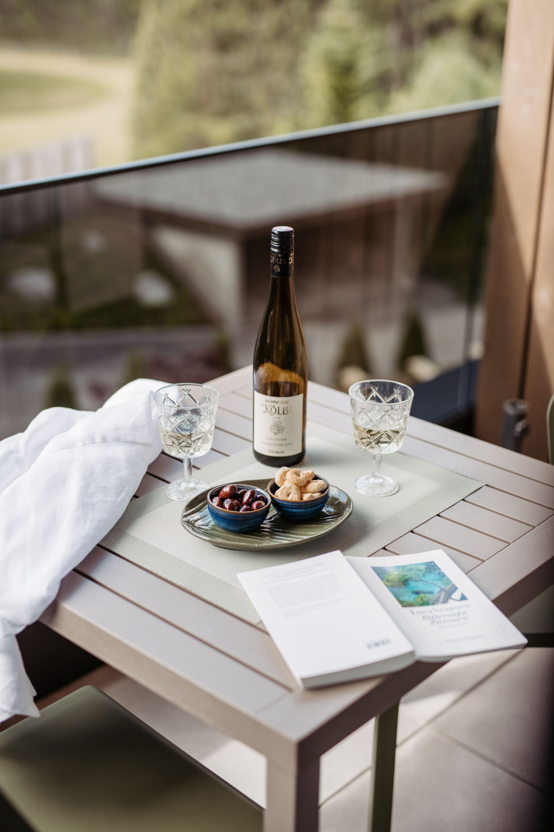 Ein Tisch mit einer Flasche, zwei Gläsern, einer Schale voll Kirschen und einem Buch auf einem Balkon.