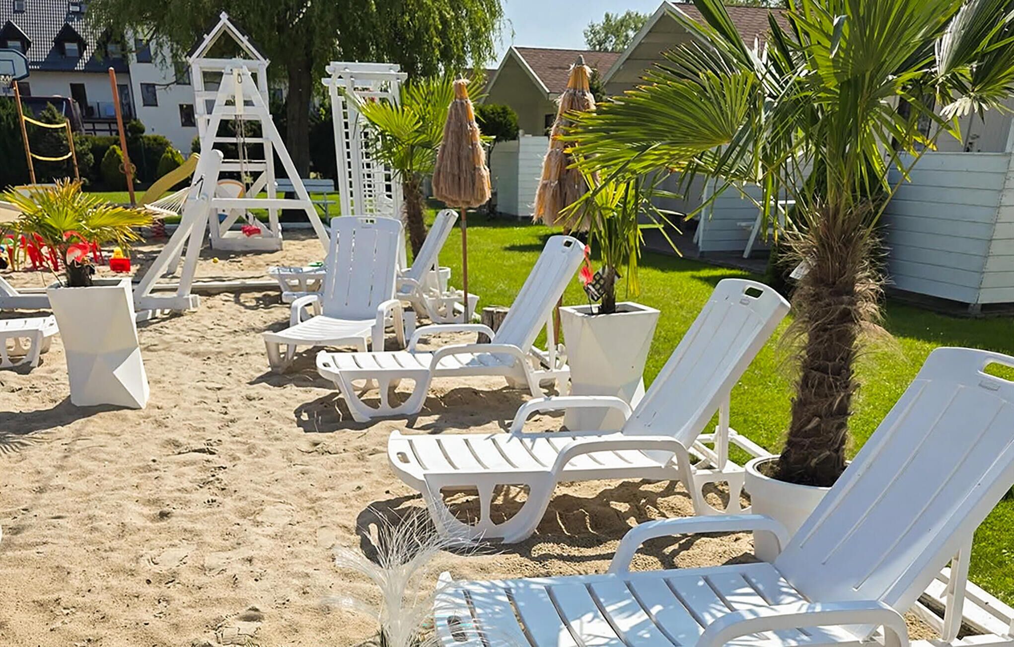 A sandy outdoor area with white lounge chairs, a small white structure, and a palm tree.