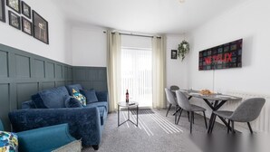 Una sala de estar moderna con un sofá azul, una mesa de comedor con sillas, una pequeña mesita auxiliar con una botella y un televisor empotrado en la pared mostrando el logotipo de Netflix.