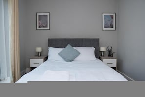 A hotel room with a large bed, two bedside tables with lamps, a grey headboard, and two framed pictures on the wall. - Spacious 4BR North Shields House Sleeps 7 Near Fish Quay Newcastle (North Shields)