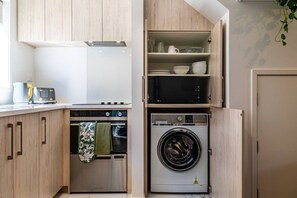 Eine kompakte Küche mit Einbauofen, Mikrowelle und Waschmaschine, Holzschränken und einem Fenster.