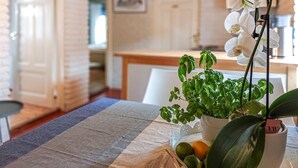 Ruokapöytä, jonka päällä on ruutukuosinen pöytäliina, ruukussa oleva basilikan taimi, ruukussa oleva orkidea ja pieni appelsiini.
