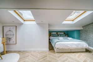 Ein Schlafzimmer mit einem Bett, zwei Dachfenstern, einem Fischgrätparkett und einer gerahmten Patentzeichnung an der Wand.