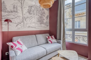 Ein Zimmer mit grauem Sofa, zwei Kissen, einer Wandlampe, einem kleinen Tisch und einem großen Fenster mit Blick auf Gebäude.