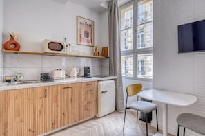 Une cuisine moderne avec des meubles en bois, un petit réfrigérateur, un four à micro-ondes et une machine à café. Une fenêtre offre une vue sur les bâtiments environnants.