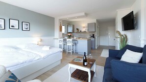 Et moderne hotelværelse med en seng, en sofa, et lille bord med en flaske og et glas, et fjernsyn og et køkkenområde i baggrunden.