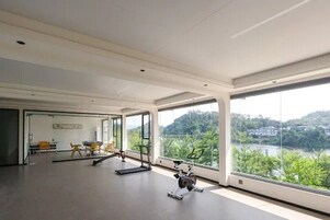 Amplia habitación con ventanas grandes, equipo de ejercicio y vistas a un lago y colinas.