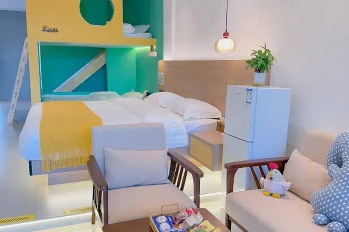 Ett hotellrum med en säng, två stolar, ett litet bord med diverse föremål, ett kylskåp och en hängande lampa.