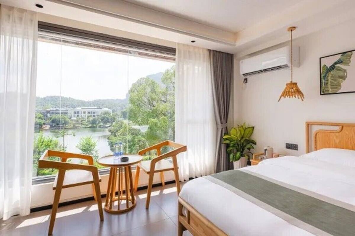 Ett hotellrum med en säng, ett litet bord med stolar, en krukväxt och utsikt över en sjö och byggnader.