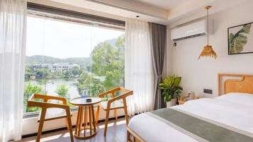 Habitación de hotel con cama, una mesita con sillas, una planta en maceta y vistas a un lago y edificios.