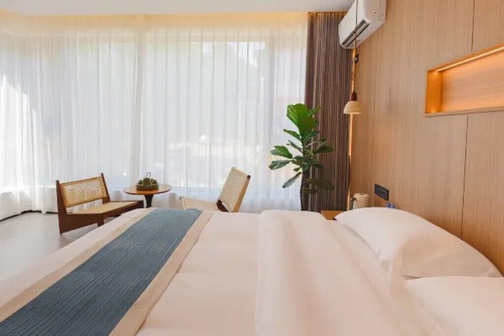A hotel room with a bed, two chairs, a small table, a potted plant, and a wall-mounted air conditioner.