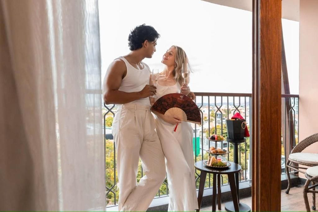 Um casal em uma varanda com vista, uma mesa pequena com comida e um ventilador.