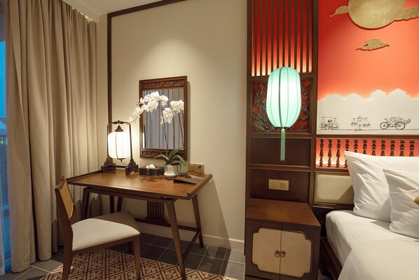 A hotel room with a wooden desk, a chair, a bed, a nightstand, a mirror, and a decorative wall with traditional artwork. - HOI AN MOON PREMIUM BOUTIQUE HOTEL & SPA (Da Nang)