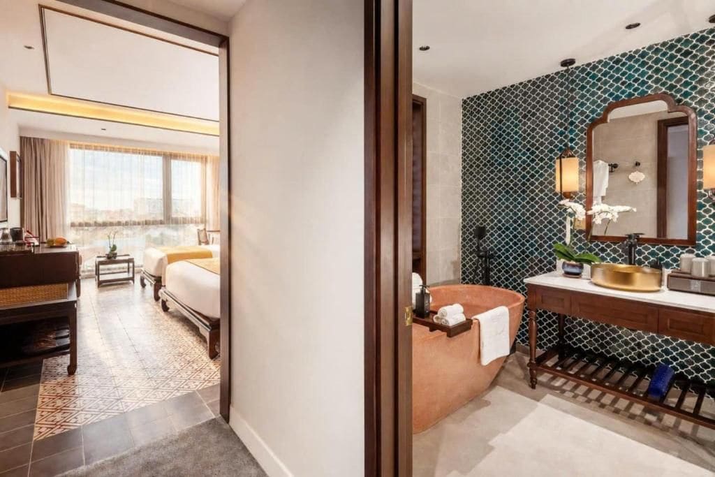 Habitación de hotel con cama, zona de estar con mesa y sillas, baño con tocador y un espejo grande, y vista a la ciudad desde la ventana.