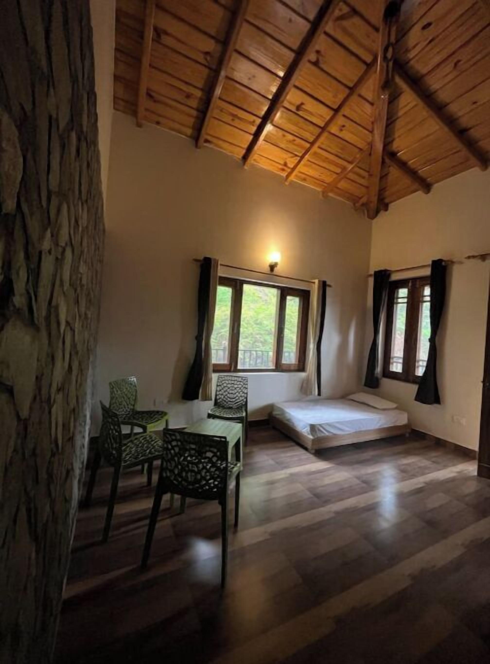 A room with a wooden ceiling, a bed, a table with chairs, and a window with curtains.
