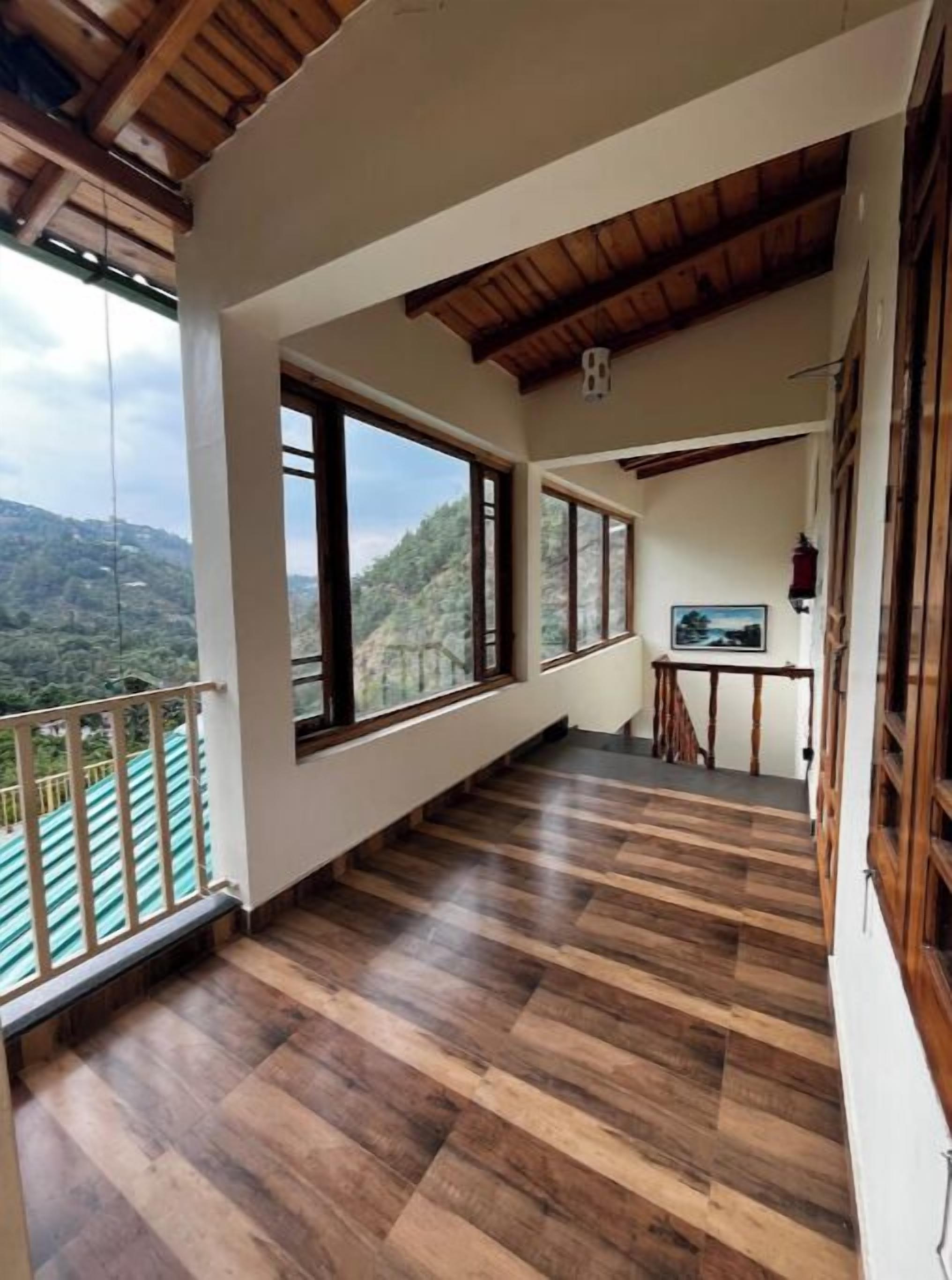Un balcon spacieux doté d’un plancher en bois, de grandes fenêtres offrant une vue sur les collines, ainsi qu’une petite table et une chaise.