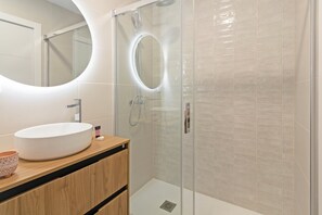 A modern bathroom with a round mirror, a white sink, and a glass-enclosed shower. - Guestready - Llovera Apartments (Valencia)