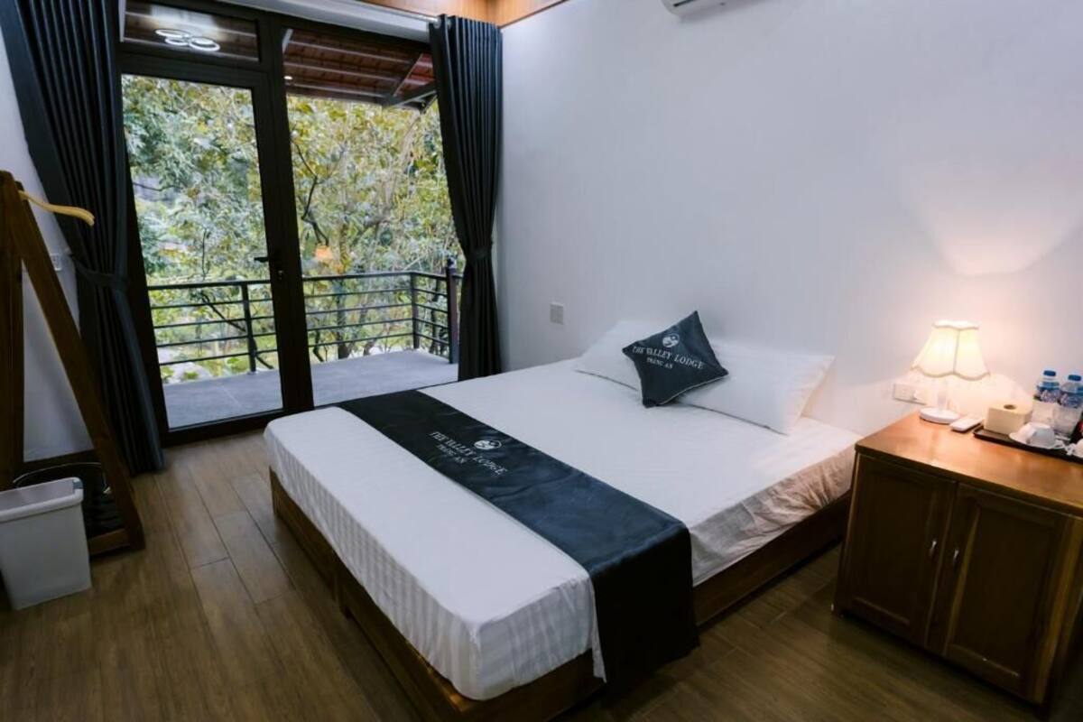 Habitación de hotel con cama, mesita de noche, lámpara, ventana con cortinas y vista a la vegetación.