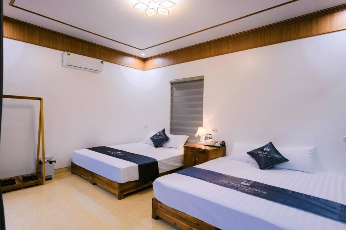 Habitación de hotel con dos camas, una mesita de noche, una lámpara, un equipo de aire acondicionado y una ventana con persianas.